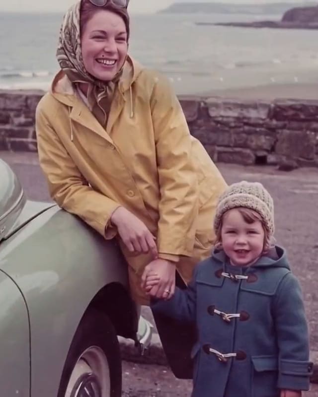 A young mother in a yellow raincoat leans on a car by the sea, holding her small son’s hand. A frame from our sample film; the people are fictional.