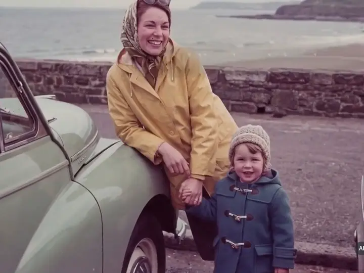 A young mother in a yellow raincoat by a car at the seafront, holding her son’s hand.