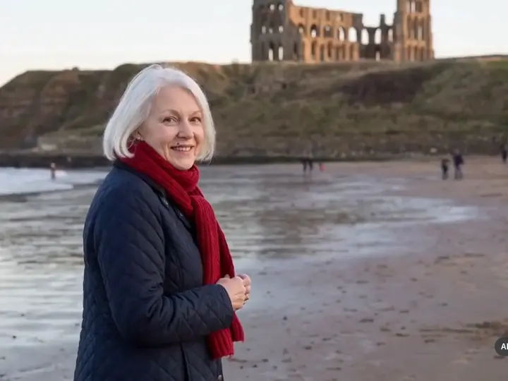 A white-haired woman in a red scarf on the beach, the abbey ruins on the cliff behind.